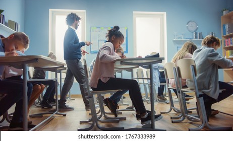 In Elementary School Class: Enthusiastic Teacher walks between Rows of Diverse Children, Explains Lesson. Group of Smart Multiethnic Kids Learning New Stuff. Side View Shot - Powered by Shutterstock