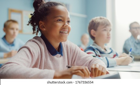In Elementary School Class: Portrait of a Brilliant Black Girl with Braces Smiles, Writes in Exercise Notebook. Junior Classroom with Diverse Group of Children Learning New Stuff - Shutterstock ID 1767336554