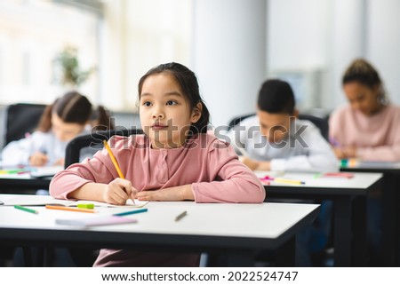 Elementary School Class. Portrait of Cute Little Asian Girl Smiling, Taking Notes And Writing in Exercise Notebook. Junior Classroom with Diverse Group of Children Learning New Stuff