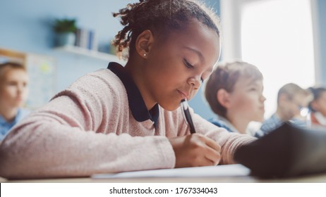 In Elementary School Classroom Black Girl Writes in Exercise Notebook, Taking Test. Junior Classroom with Diverse Group of Bright Children Working Diligently, Learning. Low Angle Side View Portrait