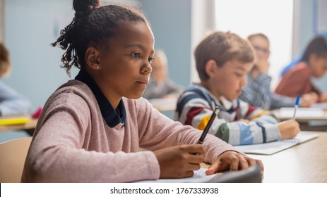 In Elementary School Classroom Brilliant Black Girl Writes in Exercise Notebook, Taking Test and Writing Exam. Junior Classroom with Diverse Group of Children Working Diligently and Learning New Stuff