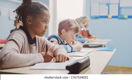 In Elementary School Classroom Brilliant Black Girl Writes In Exercise Notebook, Taking Test. Junior Classroom With Diverse Group Of Bright Children Working Diligently And Learning. Side View Portrait