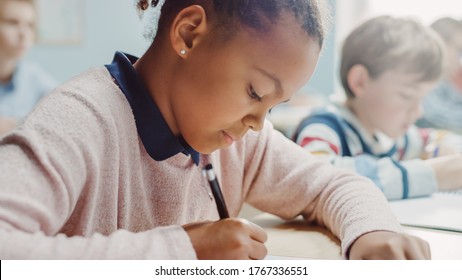 In Elementary School Classroom Girl Writes in Exercise Notebook, Taking Test. Junior Classroom with Diverse Group of Bright Children Working Diligently, Learning. Side View Portrait