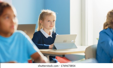 Elementary School Computer Science Class: Cute Girl Uses Digital Tablet Computer, Carefully Listening to a Teacher. Children Getting Modern Education in STEM, Playing and Learning.