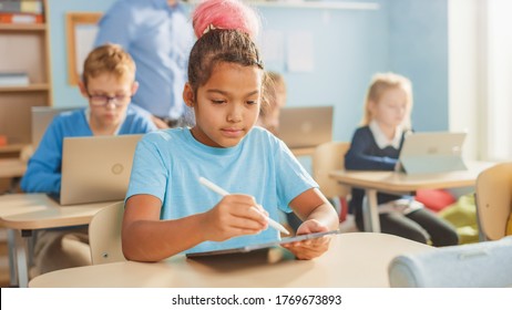 Elementary School Computer Science Class: Cute Girl Uses Digital Tablet Computer, Her Classmates work with Laptops too. Children Getting Modern Education in STEM, Playing and Learning
