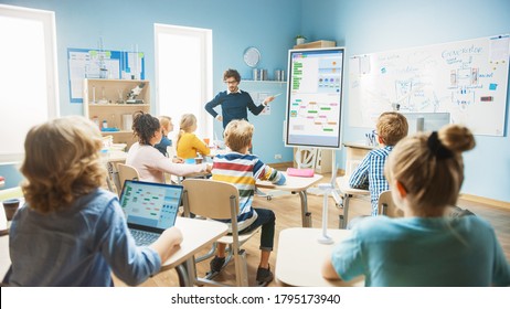 Elementary School Computer Science Teacher Uses Interactive Digital Whiteboard To Show Programming Logics To A Classroom Full Of Smart Diverse Children. Computer Class With Kids Listening