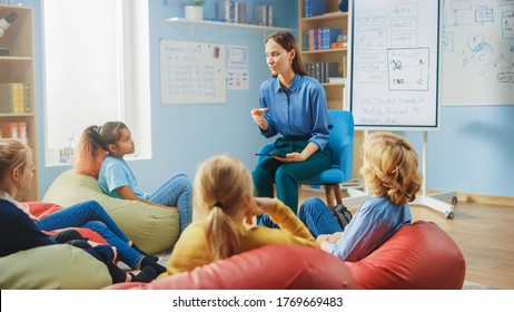 Elementary School Creativity Class: Children Sitting On The Bean Bags While Kind Teacher Explains Lesson While Using Digital Tablet Computer. Smart Children Learning In Friendly Modern Environment