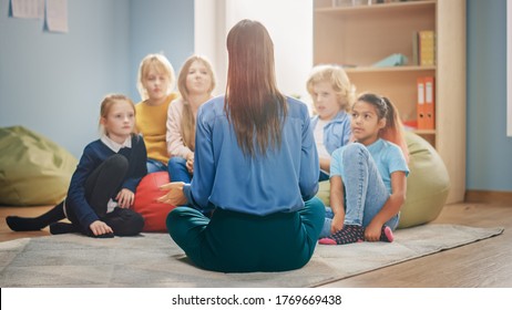 Elementary School Creativity Class: Diverse Children Sitting On Carpet While Teacher Explains Lesson And Asks Question. Learning In Modern Environment. Back View