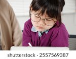 An elementary school girl with glasses studying while operating a tablet. She is receiving tutoring.