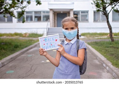 Elementary School Girl In Medical Mask Holds Picture With Back To School Message. First Offline Day With Social Distance Rules, New Normal Education, Reunion After Lockdown And Quarantine