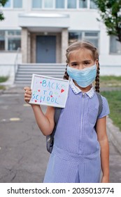 Elementary School Girl In Medical Mask Holds Picture With Back To School Message. First Offline Day With Social Distance Rules, New Normal Education, Reunion After Lockdown And Quarantine