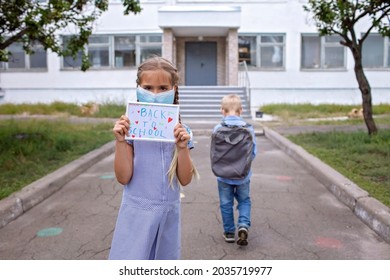 Elementary School Girl In Medical Mask Holds Picture With Back To School Message. Boy With Backpack Goes To School. First Offline Day, Study And Education, Reunion After Lockdown And Quarantine