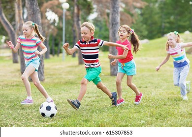 Elementary School Kids In Summer Wear Playing Soccer On Green Lawn In Park