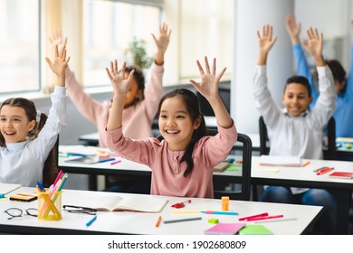 Elementary School And People Concept. Diverse Excited Group Of Emotional Happy Junior School Kids Sitting At Desks In Classroom And Raising Hands, Having Fun, Studying With Pleasure