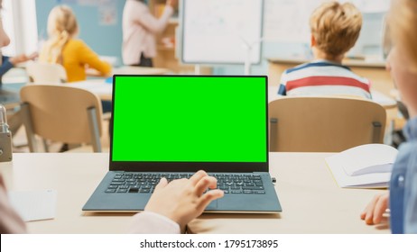 Elementary School Science Class: Little Girl Use Laptop With Green Screen Mock-up Template. In The Background School Class Full Of Diverse Smart Kids