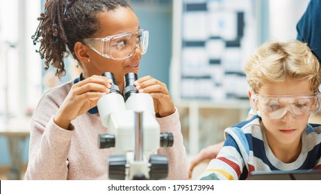 Elementary School Science Classroom: Cute Little Girl Looks Under Microscope, Boy Uses Digital Tablet Computer to Check Information on the Internet. Teacher Observes from Behind - Powered by Shutterstock
