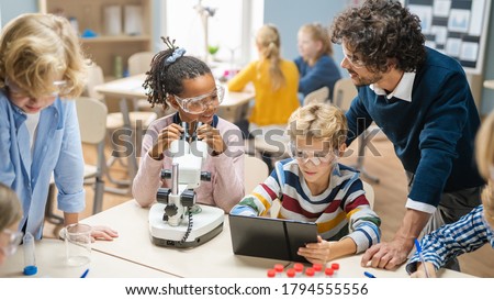 Elementary School Science Classroom: Cute Little Girl Looks Under Microscope, Boy Uses Digital Tablet Computer to Check Information on the Internet. Teacher Observes from Behind