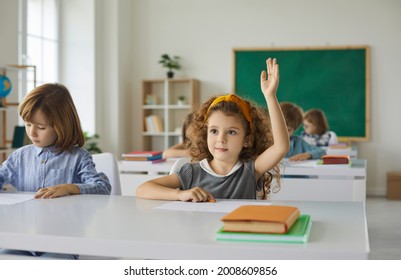 Elementary school student raises her hand, ready to answer the teacher's questions in class. Smart little curly girl is sitting at a desk next to her classmate in the classroom. Concept of education. - Powered by Shutterstock