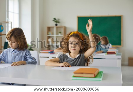 Elementary school student raises her hand, ready to answer the teacher's questions in class. Smart little curly girl is sitting at a desk next to her classmate in the classroom. Concept of education.