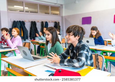 Elementary school students participating in an interactive lesson, raising hands and using laptops in a modern classroom