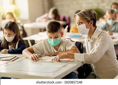 Elementary School Teacher Helping Her Student With Learning During A Class In The Classroom. They Are Wearing Protective Face Masks Due To Coronavirus Pandemic. 