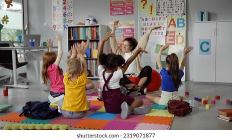 Elementary school teacher sit in class on floor with little pupils and moving hands up. Multiethnic kids with teacher in kindergarten  - Shutterstock ID 2319071877