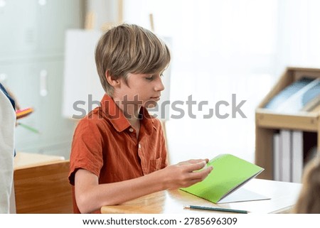 Elementary schoolboy opens green notebook at wooden desk during back to school first day, building study habit, attention skill, classroom routine, independent learning confidence in primary room.