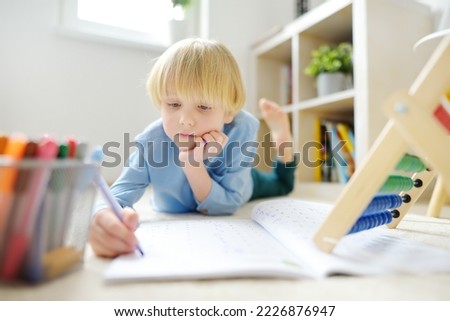 Elementary student boy doing homework on floor at home. Child learning to count, solves arithmetic examples in workbook. Math tutorial. Preparing preschooler baby for school. Education for children