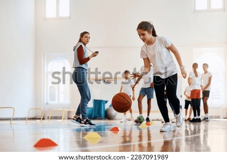 Elementary student leading a basketball during physical activity class at school gym. Her coach and friends are int he background. 
