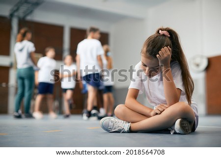Elementary student sitting away from her classmates and teacher and feeling sad during physical education class. 