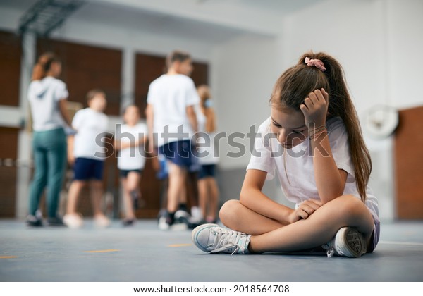 Elementary student\
sitting away from her classmates and teacher and feeling sad during\
physical education class.\
