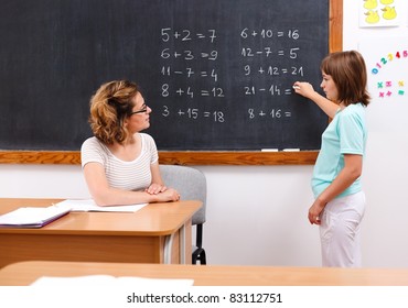 Elementary student solving equations at chalkboard while teacher looks at her