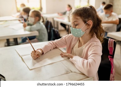 Elementary Student Writing In Her Notebook While Wearing Protective Face Mask In The Classroom During Coronavirus Epidemic. 