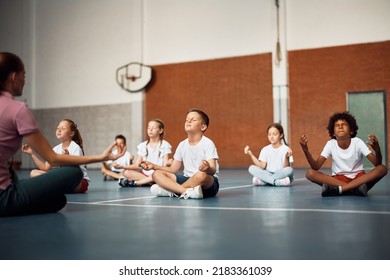 Elementary Students Meditating In Lotus Pose With Their Coach During PE Class At School Gym. Copy Space.