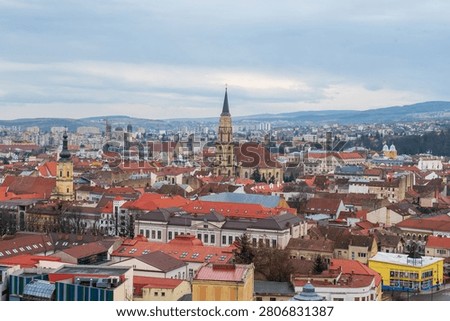 Elevated cityscape of Cluj-Napoca with red rooftops, church spires, and distant hills under an overcast spring sky in Romania.