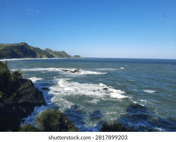 An elevated coastal view from a cliffside, capturing strong, rolling waves crashing against dark rocks. The scene includes coastal vegetation, with a palm tree on the left and a backdrop of green hills meeting the ocean.
