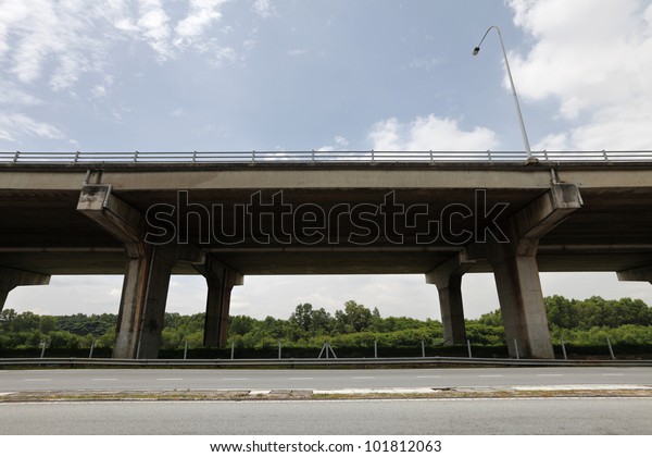 An elevated highway\
carriageway above a road along a rural green forest on a blue\
cloudy day.