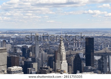 Elevated panoramic view across Midtown Manhattan featuring the iconic Chrysler Building, Queensboro Bridge and the New York City skyline beneath dramatic clouds. A classic commercial travel and architecture photograph.