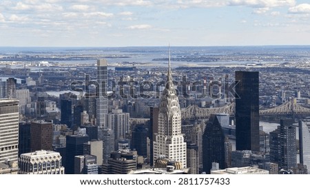 Elevated panoramic view across Midtown Manhattan featuring the iconic Chrysler Building, Queensboro Bridge and the New York City skyline beneath dramatic clouds. A classic commercial travel and architecture photograph.