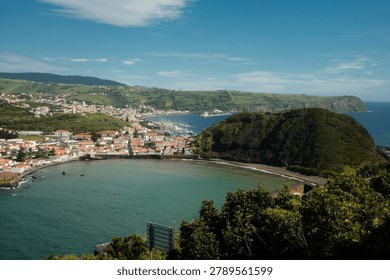 An elevated panoramic view captures the charming town of Horta on Faial Island, Azores, with its distinctive terracotta-roofed buildings curving along a turquoise bay framed by a dramatic volcanic hill and lush green cliffs extending toward the Atlantic horizon. The marina with moored sailboats, terraced hillsides, and vibrant coastal scenery reflect the unique volcanic landscape and maritime heritage of this mid-Atlantic Portuguese archipelago. Ideal for travel, tourism, and destination marketing content.