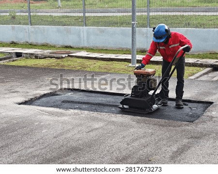 Elevated perspective view of a site worker compacting hot-mix asphalt pre-mix with a engine-driven vibratory compactor, illustrating civil infrastructure repair, pavement density compaction, and roadwork engineering.