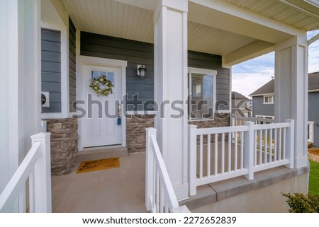 Elevated porch of a house with white front door with lockbox and wreath above the window panel. Porch with wooden railings and pillars against the wall with dark gray wood lap and stone veneer siding.