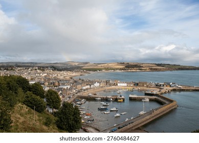 From an elevated position on top of the cliffs, looking down over the harbour and town of Stonehaven on the Scottish east cost. White fluffy clouds fil the sky above with the blue reflected in the gentle waters of the North Sea. Yachts and boats nestle between the harbour walls as the coastline stretches into the ditance.