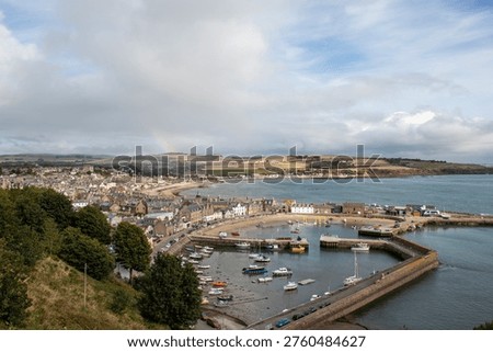 From an elevated position on top of the cliffs, looking down over the harbour and town of Stonehaven on the Scottish east cost. White fluffy clouds fil the sky above with the blue reflected in the gentle waters of the North Sea. Yachts and boats nestle between the harbour walls as the coastline stretches into the ditance.