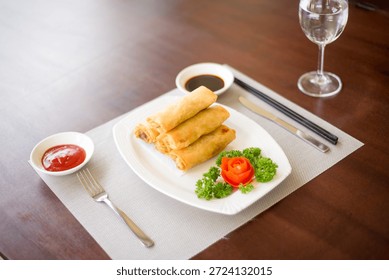 Elevated side view of a pyramid of spring rolls on a plate, accompanied by two sauces: soy sauce and a red Chinese-style sauce. The plate is placed on a mahogany table and brightly illuminated, showing a spacious and appetizing presentation from a diner’s perspective.
