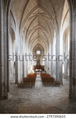 An elevated, slightly downward-angled perspective showcases the Gothic architecture inside the abbey church of Abbaye Royale Notre-Dame de Celles-sur-Belle. The high-angle framing highlights the patterned stone tile floor, towering ribbed vaults, and rows of empty wooden chairs leading symmetrically toward the distant altar.