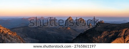 Elevated sunset view from Assekrem summit of the Atakor mountain chain. Jagged volcanic peaks glow orange against a soft gradient colorful sky in the Hoggar plateau of Tamanrasset, Algeria.