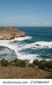 An elevated view captures a remote, rustic hut perched atop a dramatic rocky cliff, with powerful turquoise ocean waves crashing against the shore under a clear, bright sky on a sunny day.
