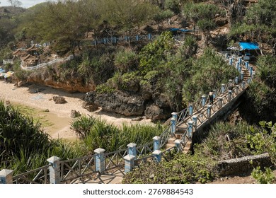 An elevated view captures a sandy beach cove featuring a blue and white wooden bridge, dense green coastal plants, and traditional huts under a bright sky.