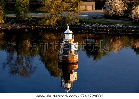 An elevated view of a charming white navigation beacon, reminiscent of a small lighthouse, standing on a sturdy stone base in the tranquil dark blue waters of Nacka. The beacon features a pointed roof, a metal staircase, and a sign displaying vessel regulations in Swedish: 'Fartyg ?ver 400 brutto 7' (Vessels over 400 gross tonnage 7) and 'Fartyg under 400 brutto 12' (Vessels under 400 gross tonnage 12). The water beautifully reflects the surrounding landscape, including lush green and blossoming trees lining the rocky shores. In the background, a road and buildings are visible under the warm, golden light of what appears to be early morning or late afternoon, creating a peaceful and picturesque scene.